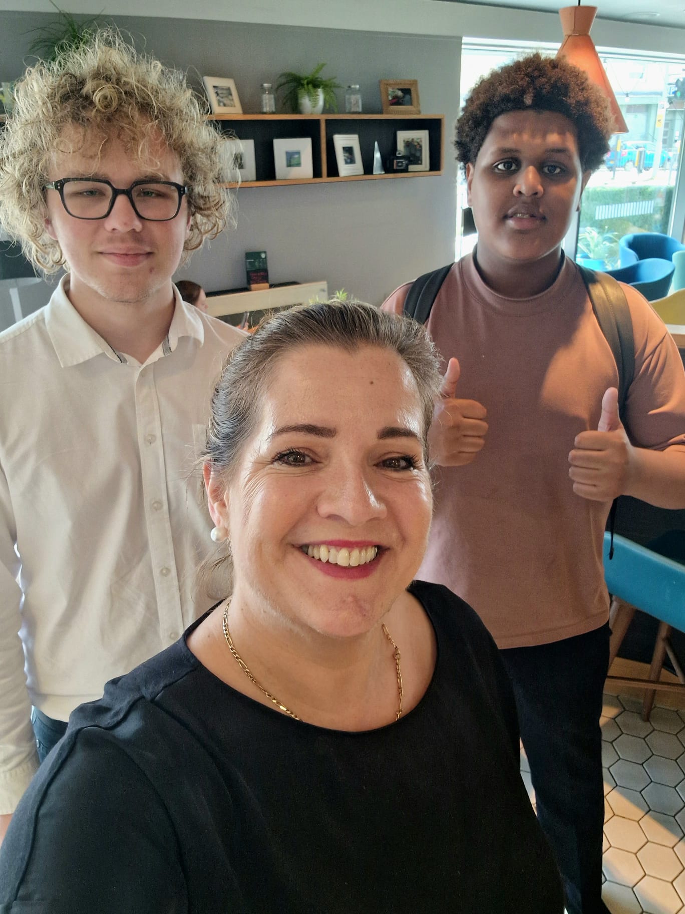 "Group selfie featuring a smiling woman in the foreground with two young men in the background. The woman, with light skin and dark hair, wears a black top and gold necklace, smiling brightly at the camera. Behind her, one young man with light skin, curly blond hair, and glasses wears a white shirt, while the other, with dark skin and short curly hair, wears a dusty rose t-shirt and gives a double thumbs-up gesture. The setting includes a light gray wall with shelves holding plants and decor, and a large window revealing an urban outdoor view with colorful furniture visible inside."
